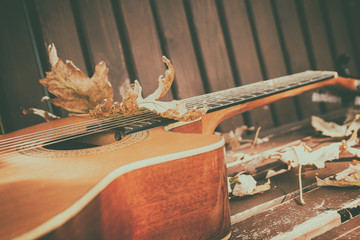 Close-up view of a fragment of acoustic classical guitar with fall maple leaf on wooden background. Autumn live concert concept. Selective soft focus.