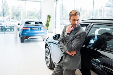 pensive bearded businessman standing near cars in car showroom