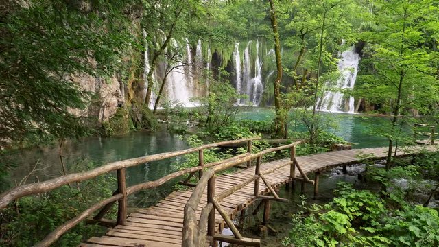 Plitvice Lakes National Park, Croatia. Walking the wooden boardwalk above a lake surrounded with rich green flora. Waterfalls in the background. UHD