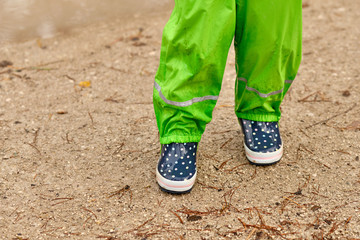 A child in green waterproof pants and spotted blue rubber boots standing on a wet gravel road in the forest on a rainy day in October in Germany