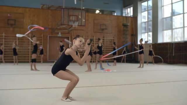Cute little girl of elementary school age rehearsing rhythmic gymnastics dance in school gym, other girls practicing in the background