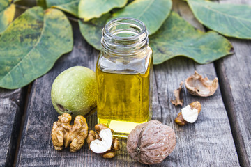 Walnut oil in a glass jar near walnuts and green leaves on a wooden table.