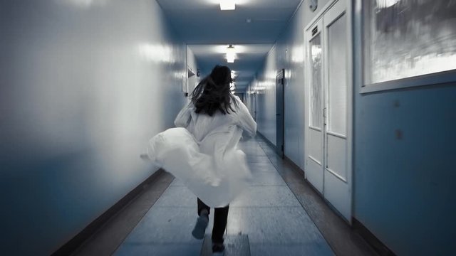 Tracking shot of zombie woman with black hair in laboratory coat walks down dark corridor goes behind door and falls,view from back. Above doorway inscription in Russian exit.