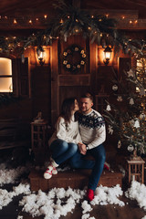 Christmas portrait of a young couple. The guy and the girl are sitting in a beautiful home. Christmas mood of lovers on background pf wooden house and decorative snow . Studio photo about Christmas