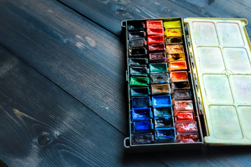 On a dark gray wooden tabletop, juicy watercolor paints, which the artist already used, are open.