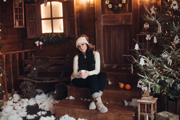 Fototapeta na wymiar Full length of dark-haired woman in warm hat and socks in white sweater sitting on wooden porch with mug of hot drink or cacao in beautifully decorated room with Christmas tree and garland. It is