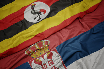 waving colorful flag of serbia and national flag of uganda.