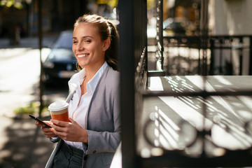 Young woman leaning on wall. Businesswoman drinking coffee and using phone.