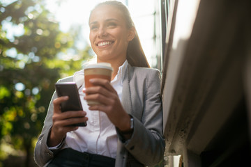 Young woman leaning on wall. Businesswoman drinking coffee and using phone.