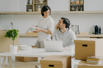 Moving into new house. Happy family couple unpack cardboard packages with dishes, pose in kitchen, relocate in modern apartment, man works on laptop, searches information for house decoration