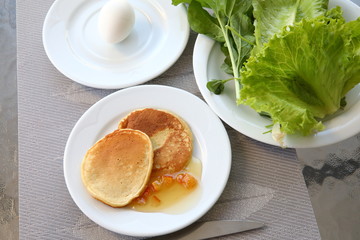 Freshly baked tasty fluffy wheat pancakes, jam with orange slices on a white plate.Bunch of fresh green lettuce and arugula leaves.Boiled egg.A satisfying start to the day.Background image close up
