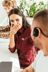 selective focus of cheerful broker covering face while laughing and looking at coworker in office