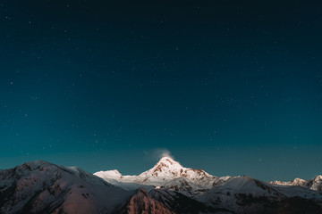 Fototapeta na wymiar Georgia. Winter Night Starry Sky With Glowing Stars Over Peak Of Mount Kazbek Covered With Snow. Beautiful Night Georgian Winter Landscape