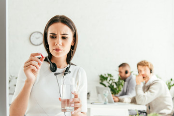 selective focus of sick broker holding pill and glass of water near coworkers in office