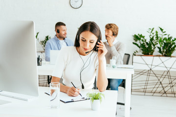 selective focus of attractive broker touching headset and writing near coworkers