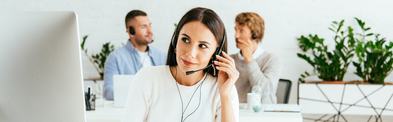 panoramic shot of attractive broker touching headset near coworkers in office