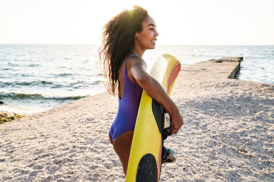Image of nice african american woman holding surfboard and smiling
