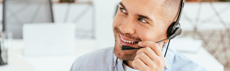 panoramic shot of happy and handsome operator in brokers agency touching headset