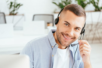selective focus of happy operator in brokers agency touching headset and looking at camera in office