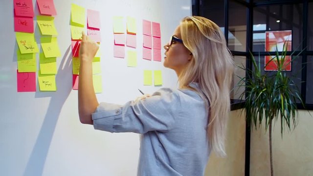Project management agile methodology, concept. A young blonde woman in glasses, gluing colored sticky notes on a magnetic Board.