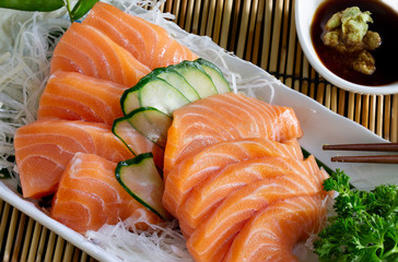 Salmon sashimi in white dish with shoyu sauce with wasabi in white bowl and chopsticks all put on bamboo mat