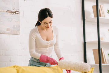 smiling housewife cleaning yellow sofa with white dusting brush