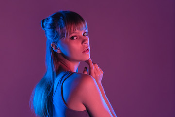 Fashion attractive young blond hair 20s fashion girl model pretty face looking at camera standing at purple studio background, sexy trendy stylish woman in pink neon party night glow concept portrait
