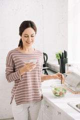 young, smiling woman using smartphone while standing at kitchen table near bowl with vegetable salad