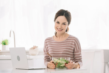Obraz na płótnie Canvas young, smiling woman looking at camera while sitting at table with bowl of vegetable salad near laptop