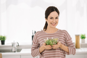 happy young woman smiling at camera while holding bowl with fresh vegetable salad