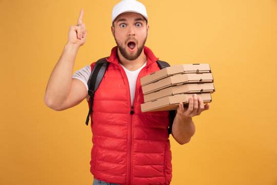 Glad delivery man has joyful expression, points up, shows where pizzeria situated, carries pile of pizza boxes, distributes product for customer, isolated on yellow wall. Advertisement and nutrition
