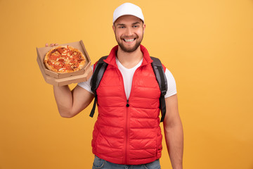 Cheerful pizza delivery man stands with carton box with pizza, waits for client, wears white cap and white tshirt with red waistcoat, smiling during transporting fast food, poses against yellow wall