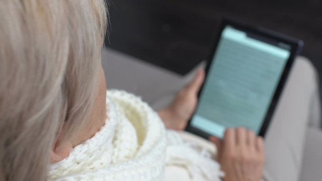 Sick senior female reading electronic book on tablet PC, seasonal epidemic