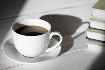 A round white cup of coffee stands on a white saucer on a white wooden table next to the shadow of the cup