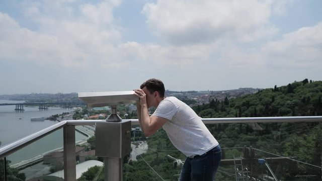 young man in white t-shirt looks through binoculars exploring landscape standing on observation deck above cable car