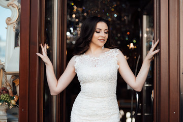 Portrait of cute brunette girl with long-haired posing on the way out on terrace in restaurant. She wears  white dress and looks happy. Beauty portrait of female face with natural skin and health hair