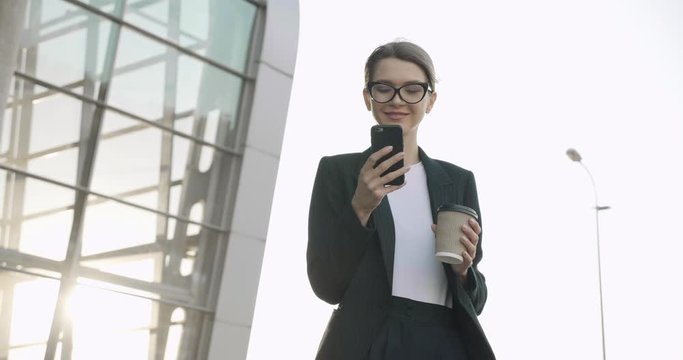 Extremely beautiful Businesswoman using her Phone, drinking Coffee take away. Young blonde Woman browsing her Smartphone near modern Office center. People. Smile. Business People. Success. Apps.