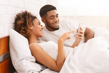 Cheerful african american couple networking on smartphone in bed