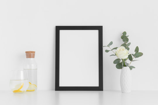Black frame mockup with a rose and eucalyptus in a vase, glass and a bottle on a white table. Portrait orientation.