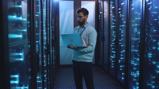 Caucasian IT engineer in high tech server room using modern laptop checking maintenance system of super computers in server rack corridor. Database workstation.