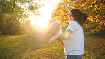Young mother spin goes around with baby child daughter kissing her and hugging on autumn park grass on bright sunset sun light among yellow leaves
