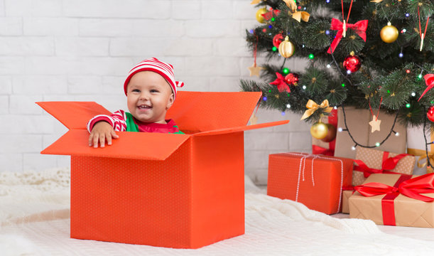 Adorable baby Santa helper sitting in present box under Christmas tree
