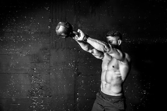 Young strong sweaty focused fit muscular man with big muscles holding heavy kettle bell for swing cross training hard core workout in the gym black and white