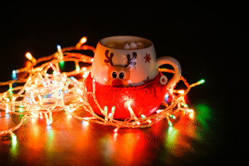 The mug is wrapped in a wool sock. A mug of cocoa and marshmallows, a garland lit next to it. Lights in the dark