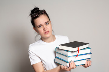 Cute, puzzled student with glasses on head holds a stack of books in hands and looks anxiously at the camera.