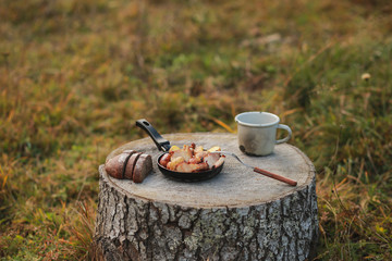 Roasted potatoes in rind with bacon, meat. Fried potatoes in a frying pan in the woods. Bread fried on a fire. Camping with food. Picnic on a stump with tea. Fried sausages. To eat in nature