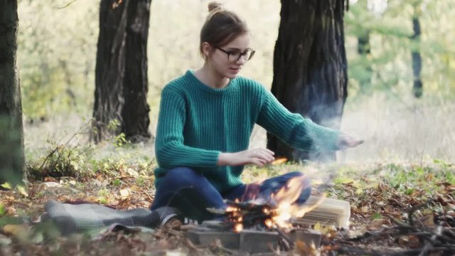 The yong caucasian woman in a warm green sweater sitting by the fire in the evening in foreat at autumn.