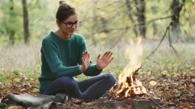The yong caucasian woman in a warm green sweater sitting by the fire in the evening in foreat at autumn.