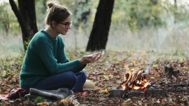 The yong caucasian woman in a warm green sweater sitting by the fire in the evening in foreat at autumn.