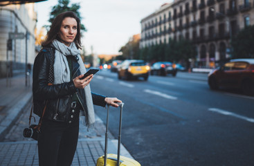 Traveler woman with suitcase calling mobile phone waiting yellow auto taxi in evening street europe...